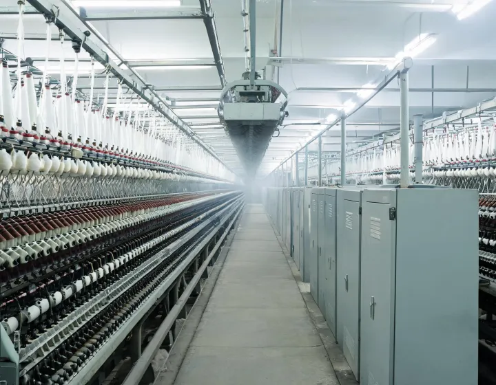 Ring spinning frames in a mill aisle with yarn packages and overhead creel